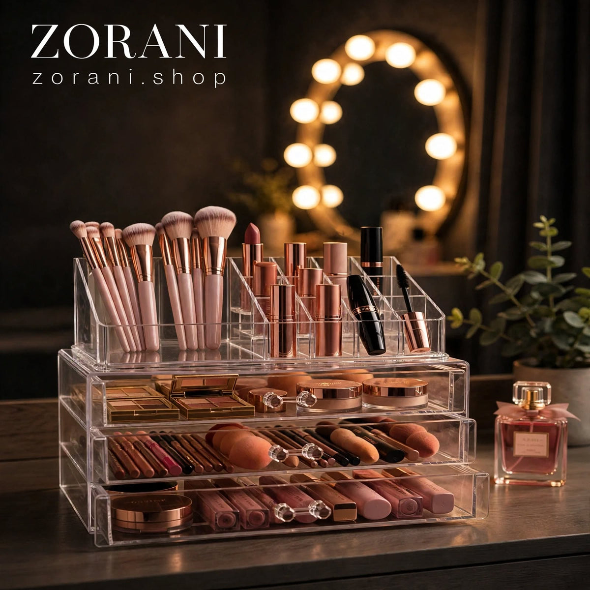 A clear acrylic makeup organizer with multiple tiers and compartments, filled with cosmetics including brushes, lipsticks, compacts, and palettes. The organizer sits on a dark tabletop in a styled vanity setting with a lit mirror, plant, and perfume bottle in the background.