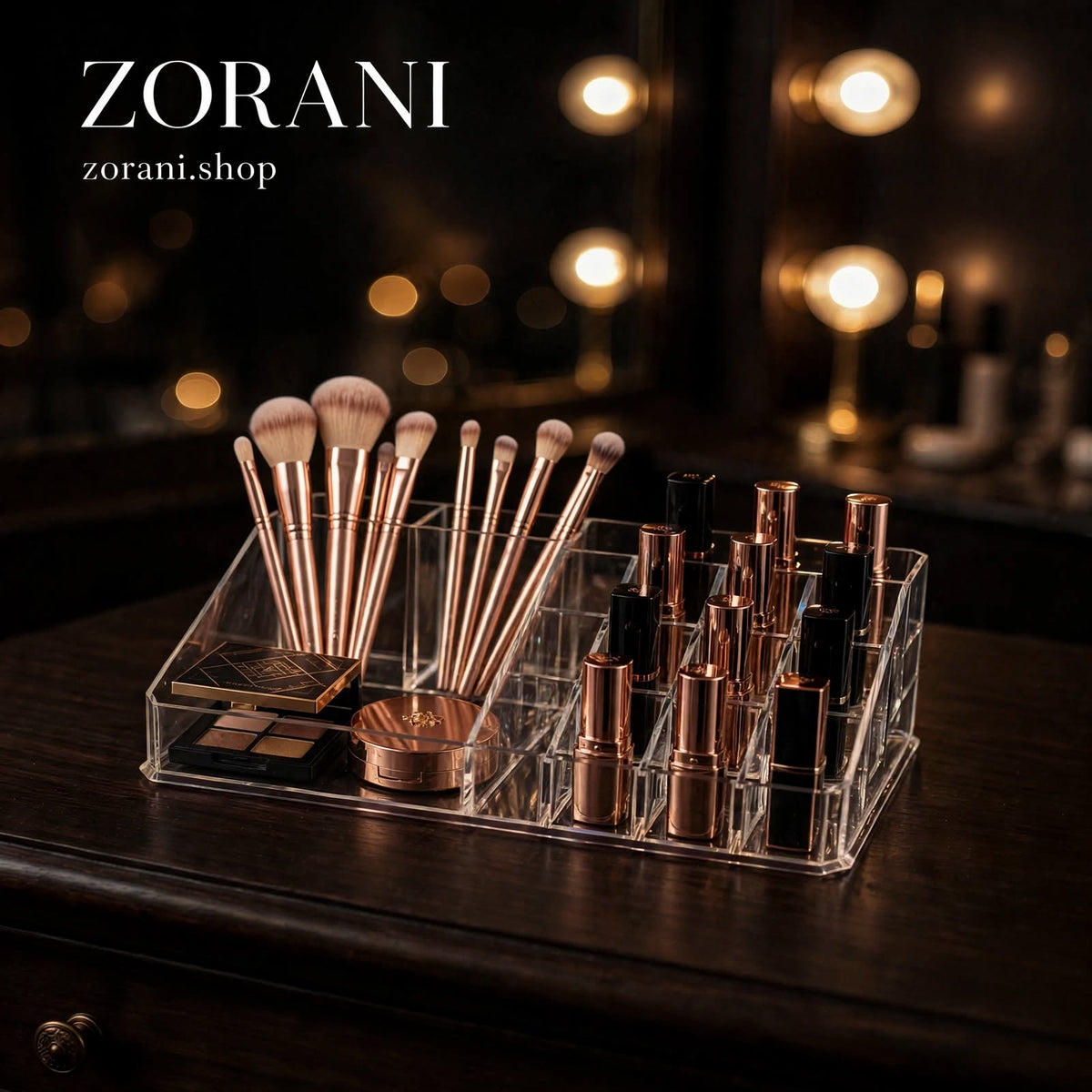 A clear acrylic makeup organizer tray holding makeup brushes, a compact palette, a powder puff, and several lipstick tubes, placed on a dark wooden vanity in a softly lit room with warm vanity lights in the background. The text 'ZORANI' and 'zorani.